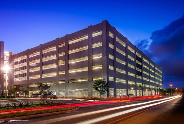 Exterior photo of the 迈阿密 Dade College Hialeah Campus parking garage. 傍晚照片与明亮的蓝色天空. 车库灯亮着. Vehicle lights create streaks on the adjacent street.